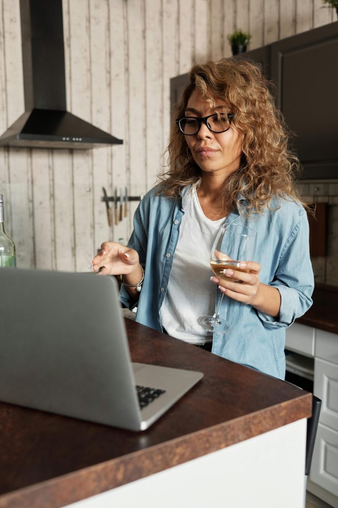 Femme aux cheveux bouclés utilisant un ordinateur portable avec un verre de vin blanc chez elle, dans une cuisine à l'ambiance chaleureuse.